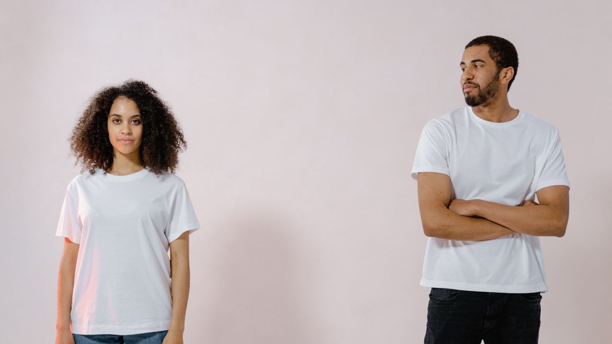 woman standing and looking straight ahead while a man looks at her. Both are wearing white t-shirts.