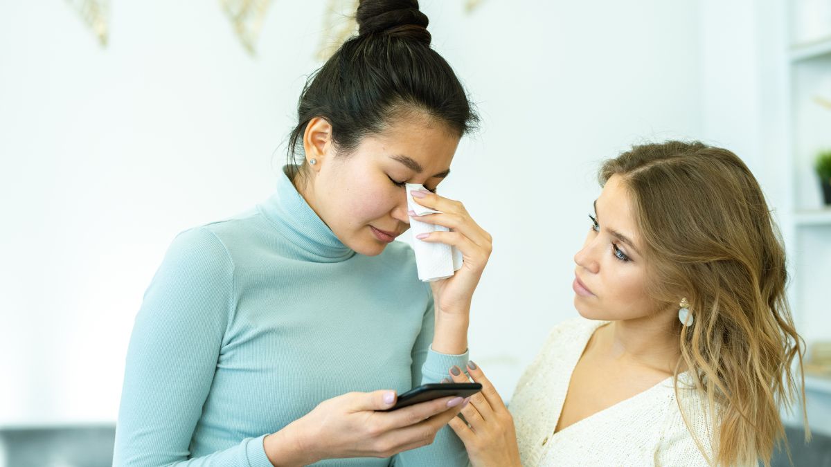 woman looking at cell phone upset while a friend consoles her