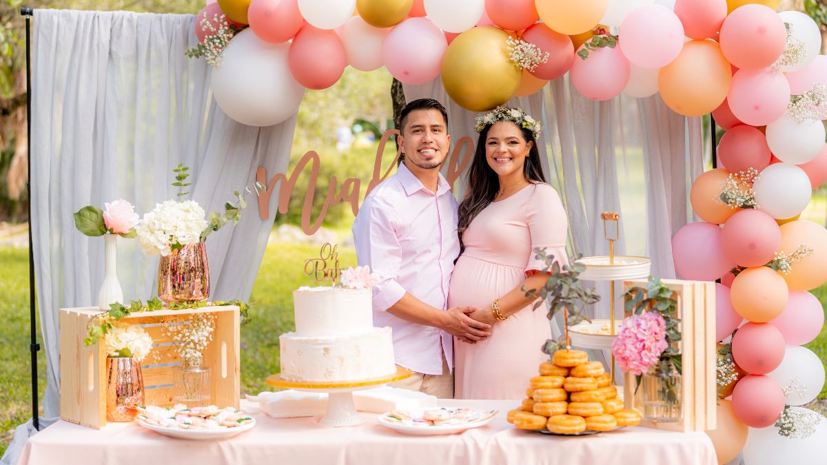 a pregnant woman and husband standing in front of decorated table and pink balloon backdrop.