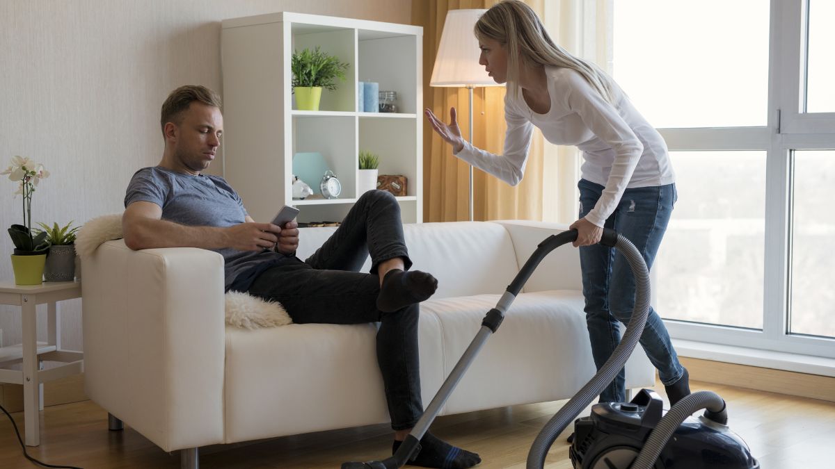 woman vacuuming while man sits on couch with cell phone