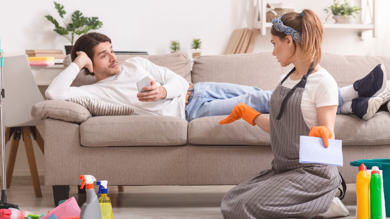 woman wearing rubber gloves and has cleaning products sitting on the ground while man lies on couch with his phone