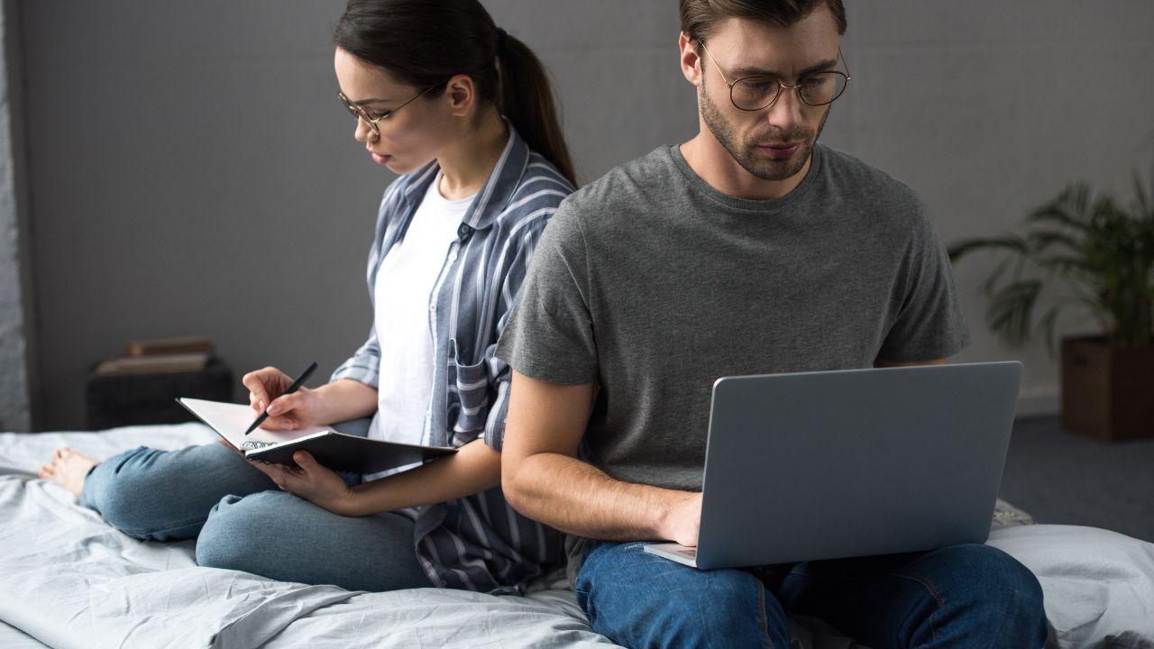 man and woman sitting on a bed. Man is working on laptop while woman writes in a notebook.