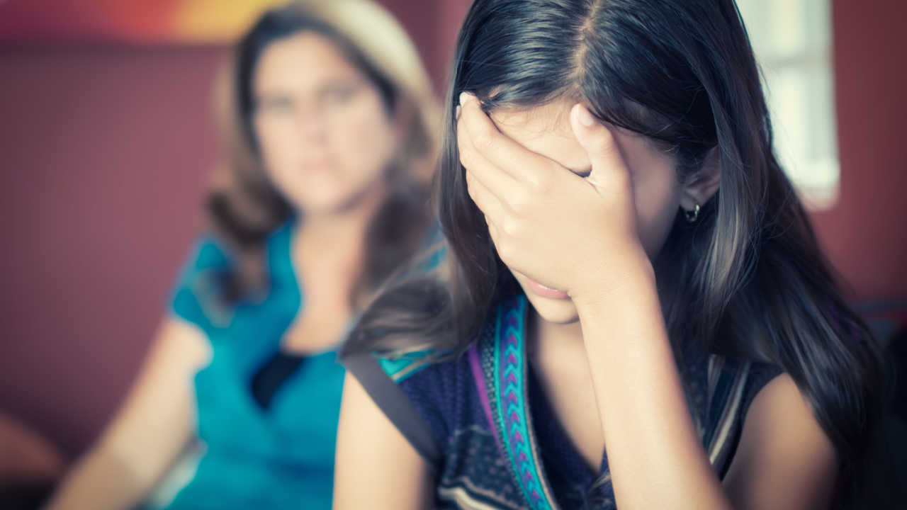 girl with hand covering face, crying while mother looks on