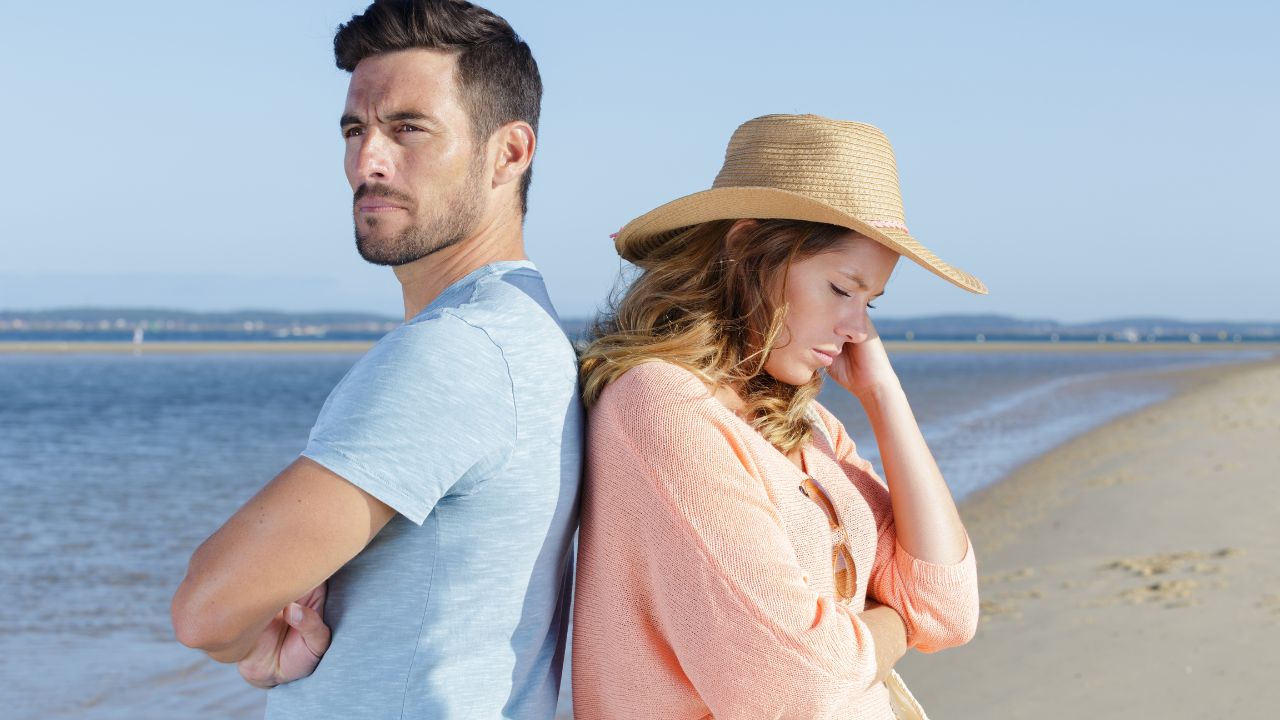 man and woman standing back to back, looking upset on the beach
