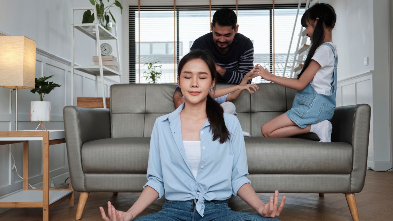 mother meditating on the floor with eyes closed while kids and husband are on the sofa behind her