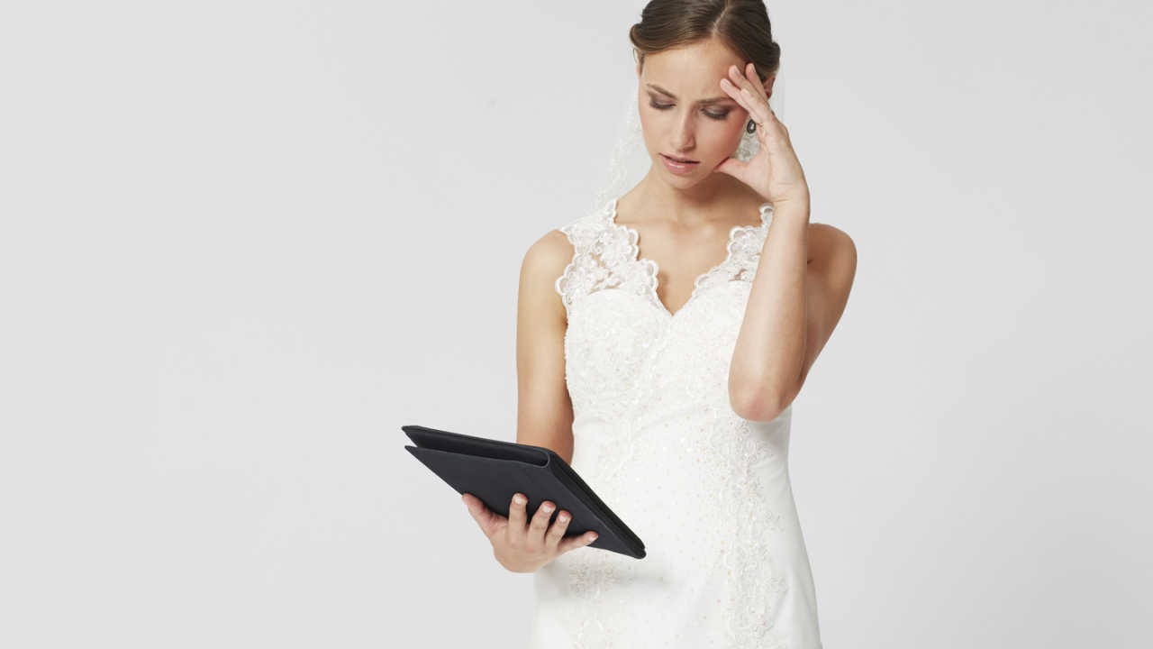 Bride holding forehead, looking at a tablet. Stressed with wedding planning.