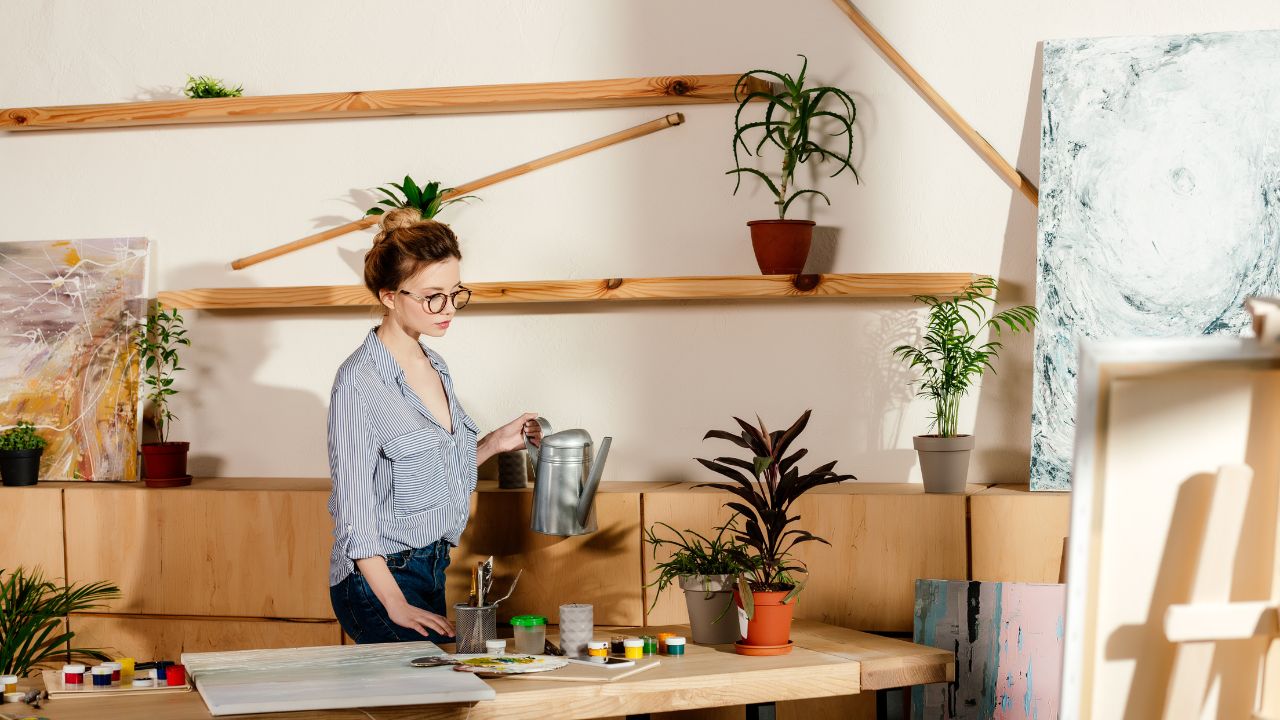 woman at work with art supplies on desk and she is holding a watering can to water her plants