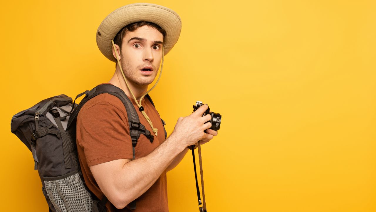 male tourist wearing a backpack and holding a camera looking shocked
