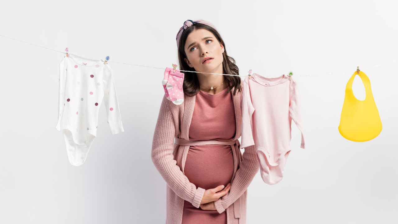 pregnant woman wearing pink clothes looking sad as she stands in front of clothes line with pink baby clothes