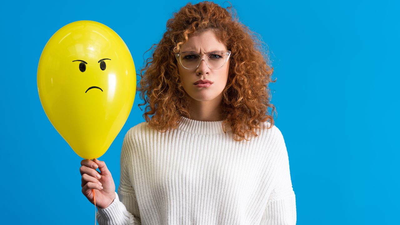 woman with red curly hair making an angry face while holding a yellow balloon with an angry face