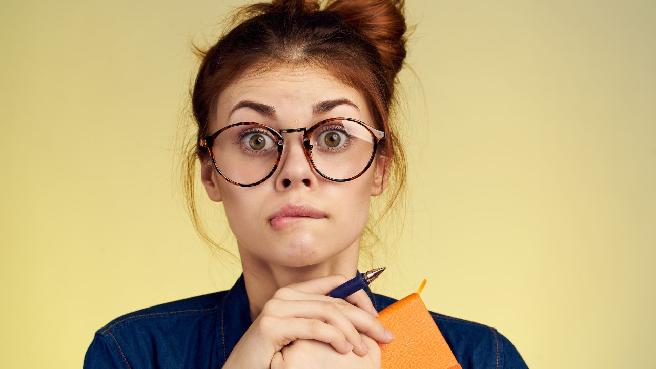 young woman wearing big glasses holding a note book and pen looking surprised, frazzled, confused