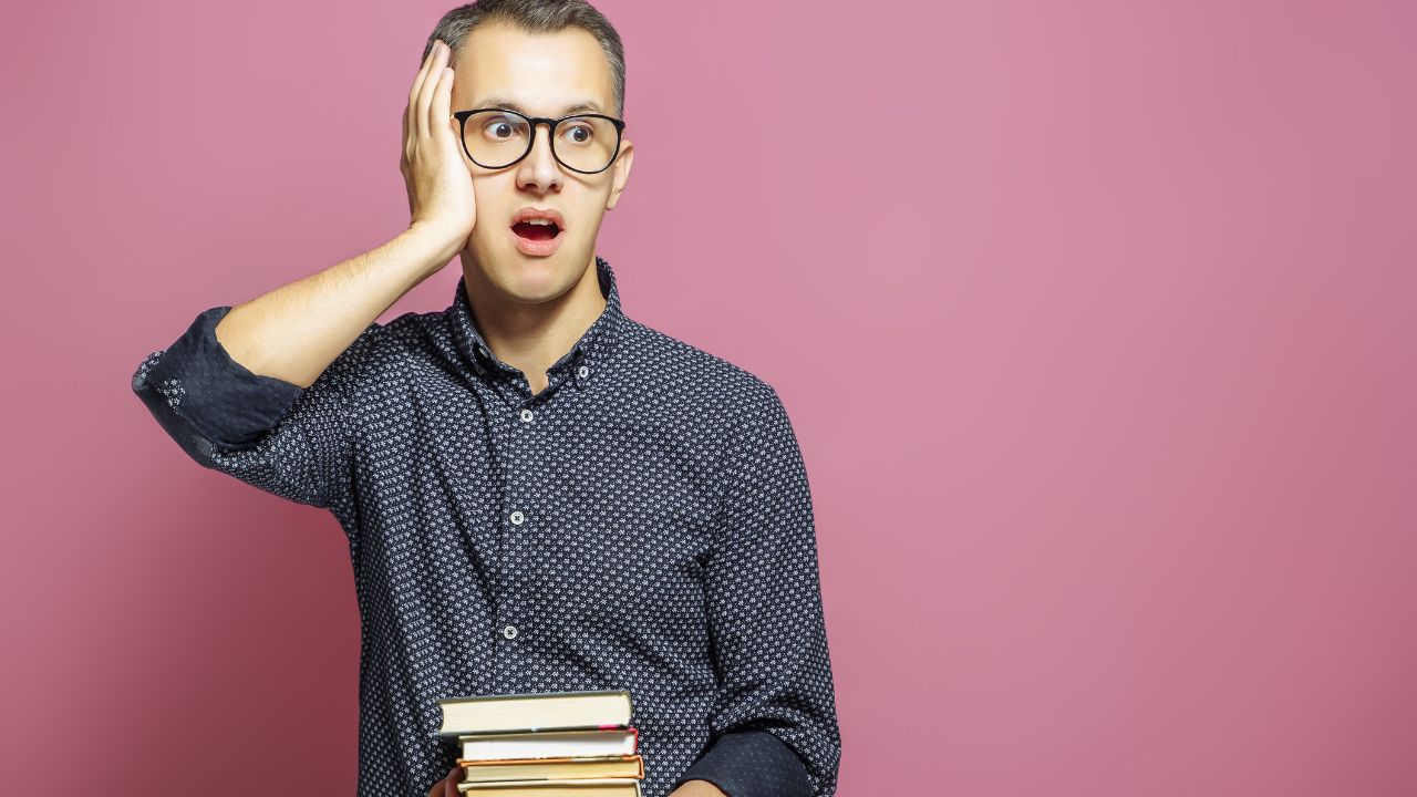 man holding a stack of books and hand on side of face looking surprised