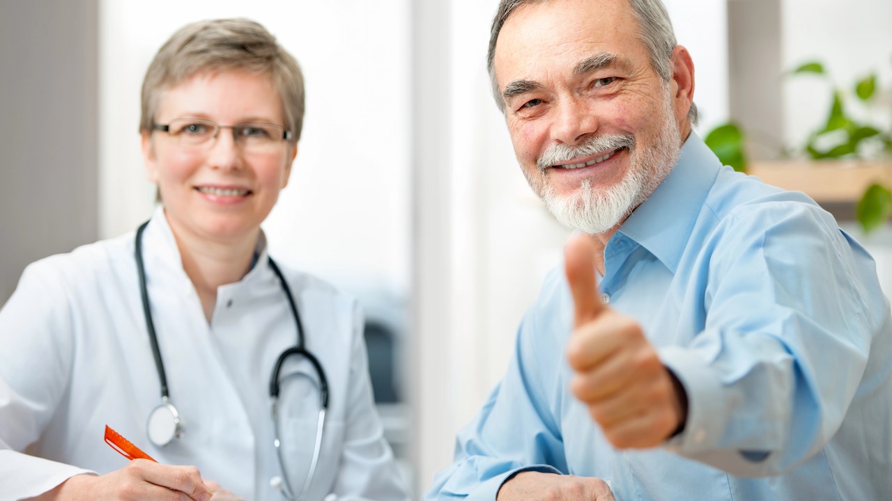 older man giving thumbs up sign after having a good doctor's appointment. He is sitting beside his female doctor.