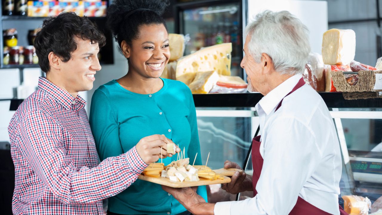 man holding cheese platter and giving free samples to a man and a woman