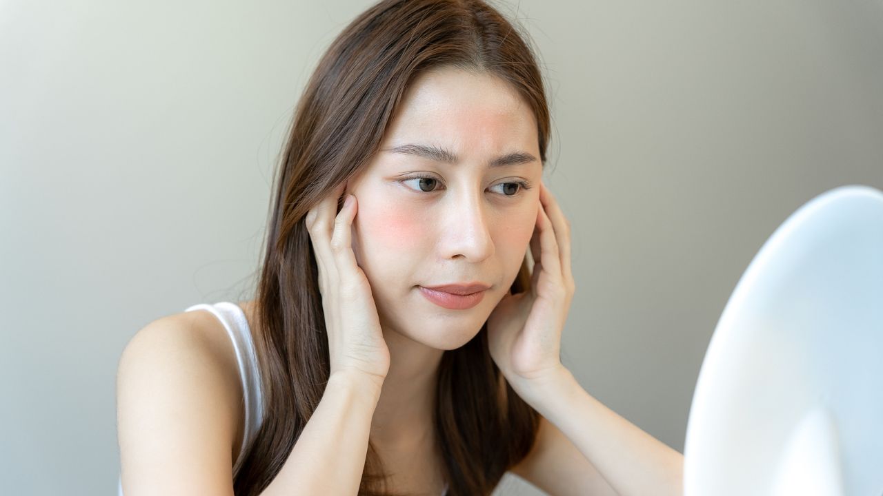 woman with rosacea looking in a mirror looking sad